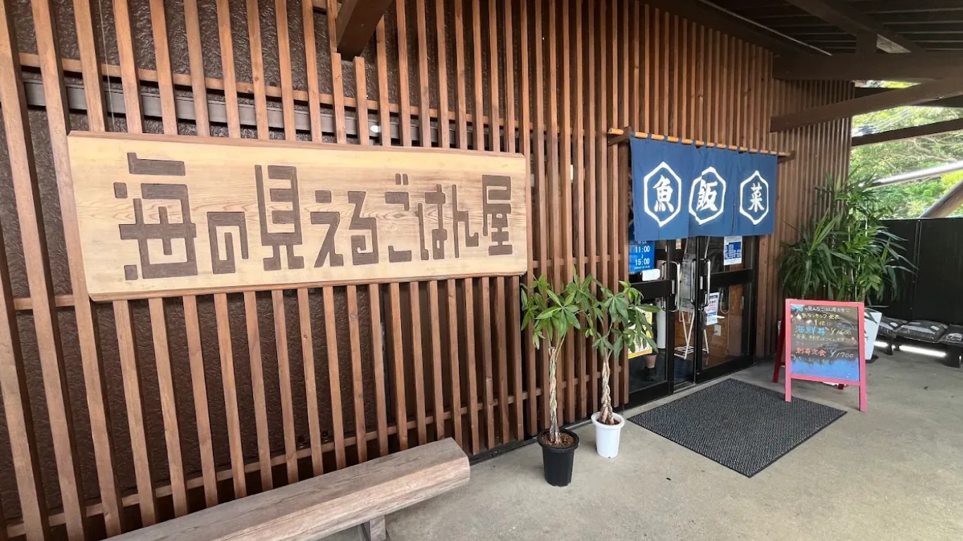 「海の見えるごはん屋」の外観。木彫りの大看板と、「魚」「飯」「菜」を白く抜いた藍の暖簾、店頭の黒板