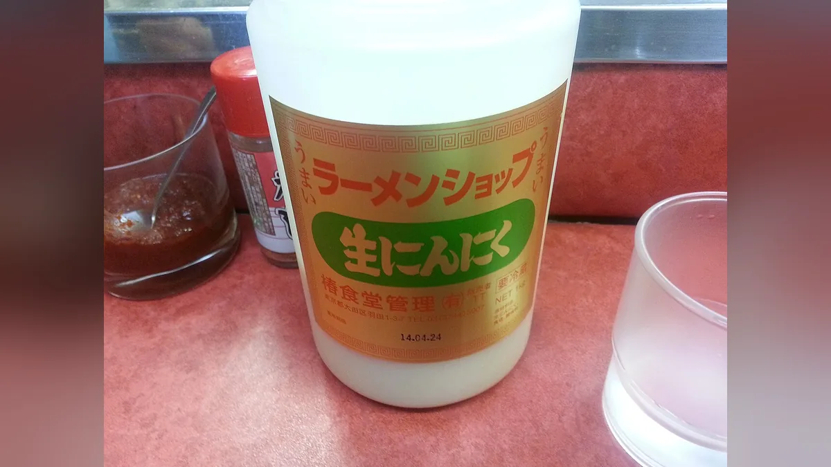 ラーメンショップの卓上に置かれた業務用「生にんにく」のボトル。ラベルに椿食堂管理有限会社の社名と大田区羽田の住所が印刷されている