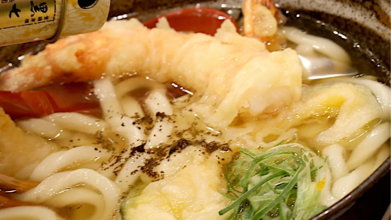 天ぷらうどんに七味唐辛子をふりかける様子